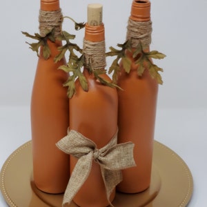 Puede incluir: Tres botellas de vidrio de color terracota con cinta de arpillera y hojas de oto&ntilde;o falsas, dispuestas en un plato de carga dorado.