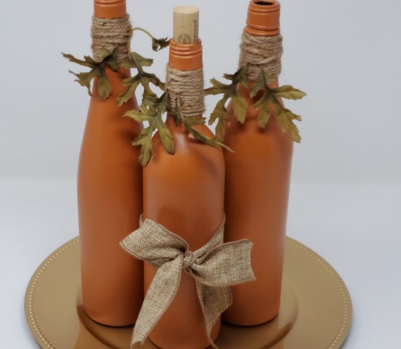 Puede incluir: Tres botellas de vidrio de color terracota con cinta de arpillera y hojas de oto&ntilde;o falsas, dispuestas en un plato de carga dorado.