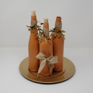 Puede incluir: Tres botellas de vidrio de color terracota con cinta de arpillera y hojas de oto&ntilde;o falsas, dispuestas en un plato de carga dorado.