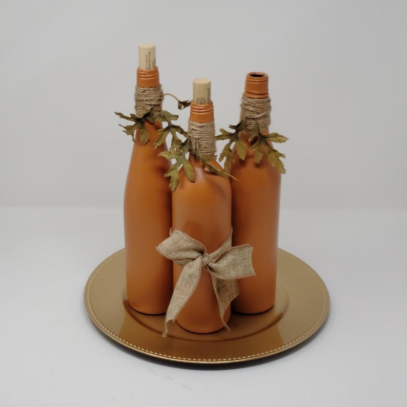 Puede incluir: Tres botellas de vidrio de color terracota con cinta de arpillera y hojas de oto&ntilde;o falsas, dispuestas en un plato de carga dorado.