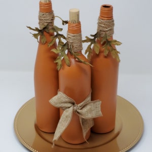 Puede incluir: Tres botellas de vidrio de color terracota con tapas de corcho, decoradas con cuerda y hojas de oto&ntilde;o falsas, est&aacute;n dispuestas en un plato dorado.