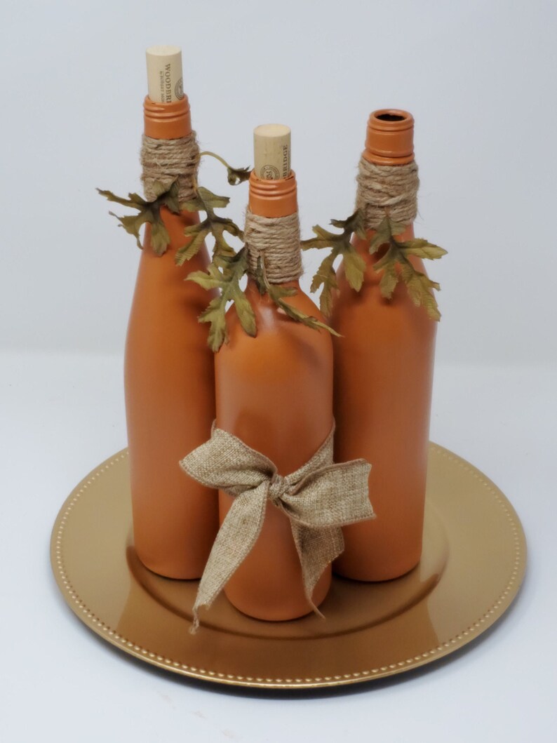 Puede incluir: Tres botellas de vidrio de color terracota con tapas de corcho, decoradas con cuerda y hojas de oto&ntilde;o falsas, est&aacute;n dispuestas en un plato dorado.