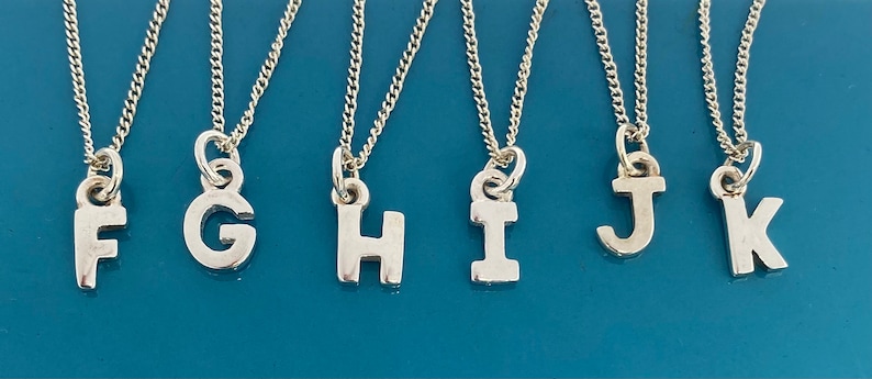 K&ouml;nnte beinhalten: Eine Auswahl an silberfarbenen Initialen-Halsketten, jede mit einem anderen Buchstaben von F bis K. Die Buchstaben sind in einer einfachen, serifenlosen Schriftart gehalten und h&auml;ngen an zarten Silberketten. Die Halsketten werden vor einem tealblauen Hintergrund pr&auml;sentiert.