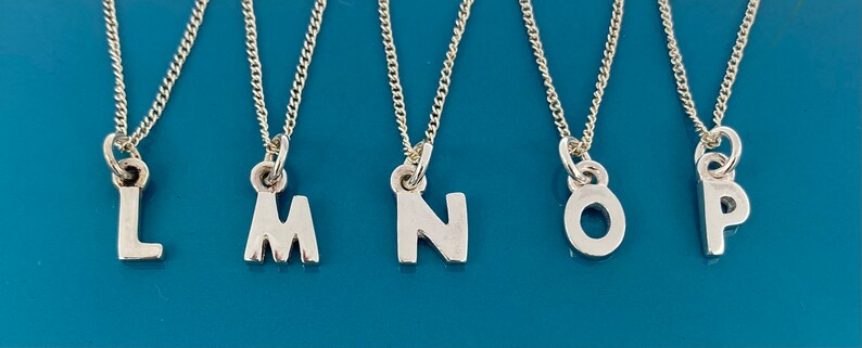 K&ouml;nnte beinhalten: F&uuml;nf silberne Halsketten mit Initialen, jeweils mit einem anderen Buchstaben: L, M, N, O und P. Die Buchstaben sind in einer einfachen Schriftart gehalten und h&auml;ngen an feinen Silberketten. Der Hintergrund ist in einem T&uuml;rkisblau gehalten.