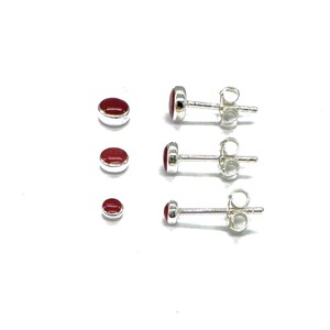May include: Three pairs of silver stud earrings with red oval-shaped stones. The earrings are arranged in order of size, with the smallest pair at the bottom.