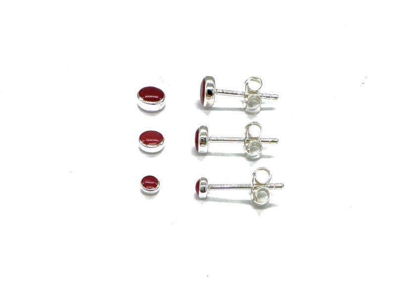 May include: Three pairs of silver stud earrings with red oval-shaped stones. The earrings are arranged in order of size, with the smallest pair at the bottom.