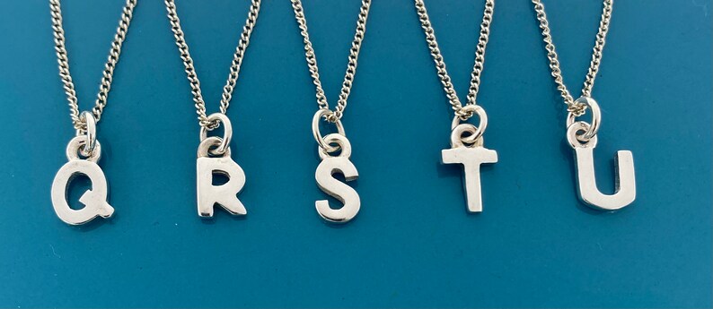 K&ouml;nnte beinhalten: F&uuml;nf silberfarbene Halsketten mit Buchstabenanh&auml;ngern. Die Buchstaben Q, R, S, T und U sind an filigranen Ketten zu sehen. Die Anh&auml;nger sind in einer serifenlosen Schriftart auf blauem Hintergrund.