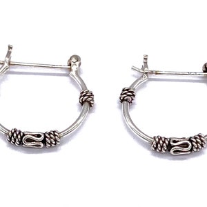 May include: A pair of silver hoop earrings with a decorative design. The hoops are small and have a twisted wire design with a small, decorative charm.