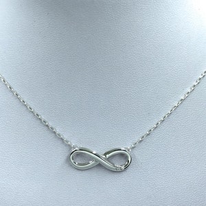 Könnte beinhalten: Eine silberfarbene Halskette mit einem Anhänger in Form eines Unendlichkeitszeichens. Der Anhänger hat einen kleinen, klaren Stein, der in einer Schlaufe eingelassen ist. Die Kette ist filigran und besteht aus kleinen, miteinander verbundenen Gliedern.