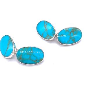 Op de afbeelding: Een paar zilveren manchetknopen met ovale turquoise stenen. De stenen hebben een blauw en groen marmerpatroon.