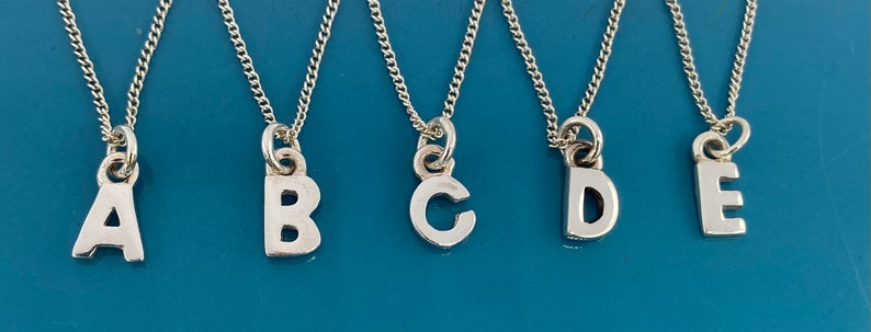 K&ouml;nnte beinhalten: F&uuml;nf silberfarbene Halsketten mit Initialen, jede mit einem anderen Buchstaben: A, B, C, D und E. Die Buchstaben sind in einer einfachen, blockartigen Schriftart gehalten und h&auml;ngen an zarten Silberketten. Der Hintergrund ist einfarbig mittelblau.
