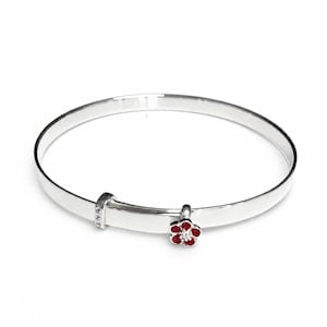 Puede incluir: Una pulsera de tipo brazalete en plata pulida con una banda lisa y redondeada. La pulsera incluye un pequeño dije colgante con forma de flor de cinco pétalos con esmalte rojo y una piedra clara en el centro, además de una pequeña cuenta plateada.