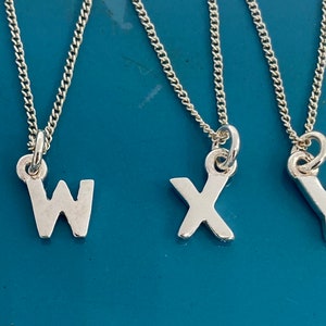 K&ouml;nnte beinhalten: F&uuml;nf silberfarbene Halsketten mit den Buchstaben V, W, X, Y und Z. Jeder Buchstabenanh&auml;nger h&auml;ngt an einer feinen Kette. Die Halsketten werden vor einem t&uuml;rkisfarbenen Hintergrund pr&auml;sentiert.