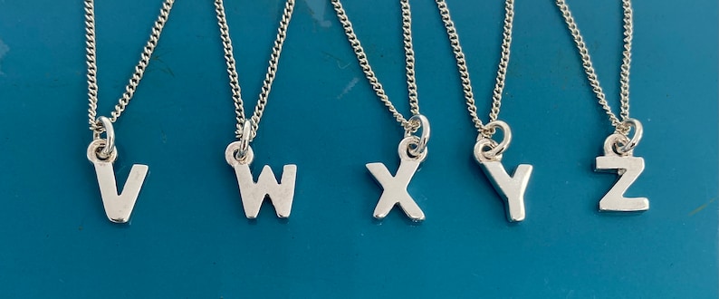 K&ouml;nnte beinhalten: F&uuml;nf silberfarbene Halsketten mit den Buchstaben V, W, X, Y und Z. Jeder Buchstabenanh&auml;nger h&auml;ngt an einer feinen Kette. Die Halsketten werden vor einem t&uuml;rkisfarbenen Hintergrund pr&auml;sentiert.