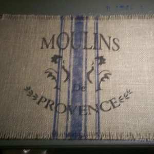May include: Rectangular burlap placemat with frayed edges. The placemat features the text "MOULINS De PROVENCE" in black, with blue vertical stripes and floral accents. The overall color scheme is neutral with a rustic aesthetic.
