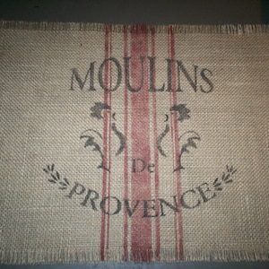 May include: A rectangular burlap placemat with frayed edges. The mat features the words "MOULINS De PROVENCE" in black, with red vertical stripes and decorative rooster and floral designs.