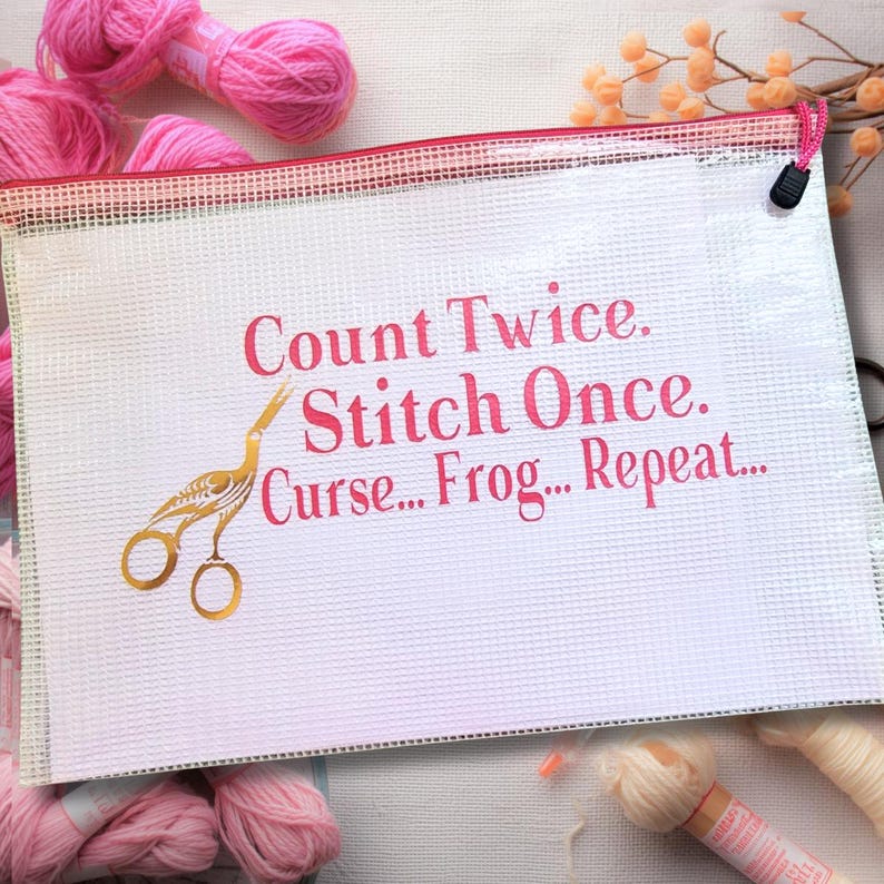 May include: White zippered craft project bag with pink text that reads "Count Twice. Stitch Once. Curse... Frog... Repeat..." and a gold scissors graphic. Pink yarn skeins are visible in the background.