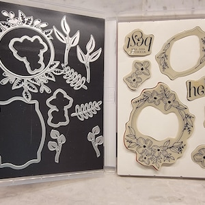 May include: A set of clear plastic storage cases containing metal and rubber stamps. The metal stamps are in a black case and feature floral wreaths, leaves, and other designs. The rubber stamps are in a white case and feature floral wreaths, leaves, and words like "hello", "love", and "&" with the text "I'll do anything for you".