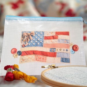 May include: A clear zippered pouch with a blue trim, featuring a patchwork American flag design. Decorative buttons and embroidery floss are visible. The flag design incorporates various patterns and textures.