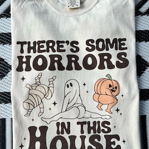 May include: A beige t-shirt with a brown graphic print that says "There's some horrors in this house". The graphic features a mummy, a ghost, and a pumpkin in a playful pose.