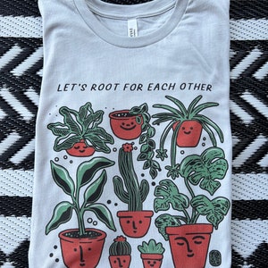 May include: A light blue t-shirt with a graphic of potted plants and the text "Let's root for each other & watch each other grow".