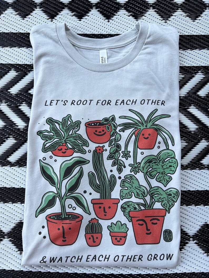 May include: A light blue t-shirt with a graphic of potted plants and the text "Let's root for each other & watch each other grow".