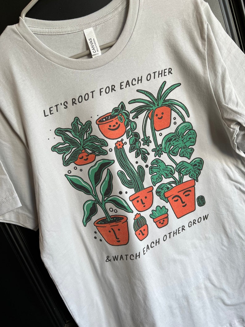 May include: A light gray t-shirt with a graphic of potted plants and the text "Let's root for each other & watch each other grow".