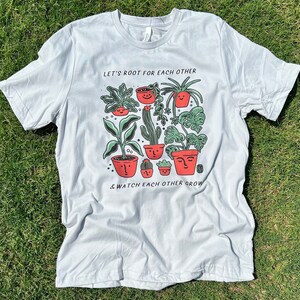May include: A light gray t-shirt with a graphic of potted plants with faces. The text "Let's root for each other & watch each other grow" is printed on the shirt.