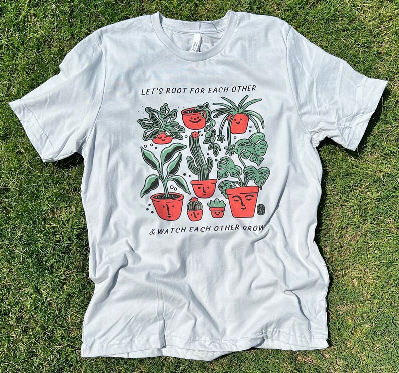 May include: A light gray t-shirt with a graphic of potted plants with faces. The text "Let's root for each other & watch each other grow" is printed on the shirt.