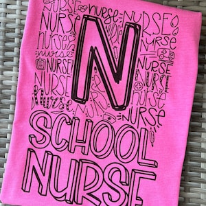 A pink t-shirt with the words 'SCHOOL NURSE' printed in black. The word 'NURSE' is repeated multiple times in a stylized font to form the letter 'N'.