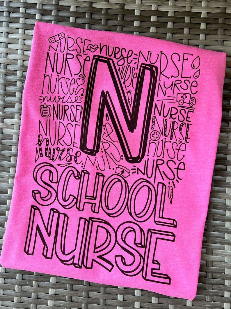 A pink t-shirt with the words 'SCHOOL NURSE' printed in black. The word 'NURSE' is repeated multiple times in a stylized font to form the letter 'N'.
