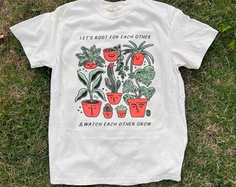 May include: White t-shirt with a green and red illustration of potted plants with faces. The text "Let's root for each other & watch each other grow" is printed on the shirt.