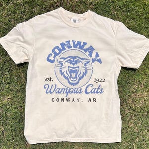 May include: A cream-colored t-shirt with a blue and white graphic of a snarling cat with the text "Conway est. 1922 Wampus Cats Conway, AR".