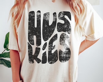 May include: Cream-colored t-shirt with the word "HUSKIES" in large, distressed black letters. The shirt has short sleeves and a relaxed fit. The text is in a bold, retro font, giving it a vintage feel. The shirt is made of a soft material.