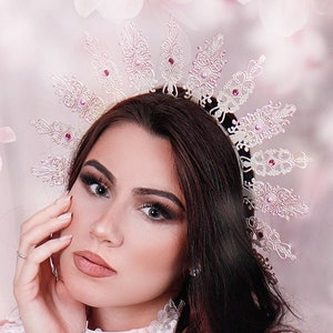 Könnte beinhalten: Eine Frau in einem weißen Spitzen- und Federkostüm mit rosa Blumen. Sie trägt eine Krone mit einem Sternendesign und hat langes dunkles Haar. Der Hintergrund ist zartrosa mit weißen Blumen.