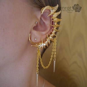May include: Gold-colored ear cuff featuring a skeletal design with multiple points and dangling chains. The cuff wraps around the ear and includes a small hoop earring. The design is intricate and detailed, with a polished finish.