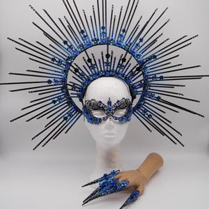 Könnte beinhalten: Ein himmlisches Ensemble mit einem schwarzen und blauen Sonnenkranz, einer passenden Augenmaske und einer Hand mit langen, spitzen blauen Nägeln. Krone und Maske sind mit blauen Verzierungen versehen. Die Artikel werden vor weißem Hintergrund präsentiert.