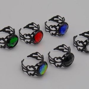 Könnte beinhalten: Eine Sammlung verstellbarer Ringe mit schwarzen Filigranbändern. Jeder Ring hat einen großen, ovalen Edelstein in verschiedenen Farben, darunter Grün, Rot, Blau, Schwarz, Weiß und ein regenbogenfarbener Schimmer. Die Ringe werden vor hellem Hintergrund gezeigt.