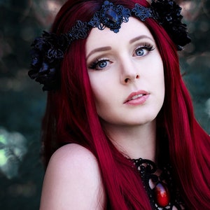 May include: A woman with long, bright red hair wears a black lace crown with a large black flower on the side. She is wearing a black lace choker with a large red gemstone pendant.
