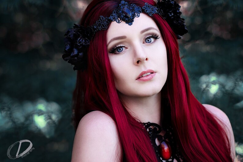 May include: A woman with long, bright red hair wears a black lace crown with a large black flower on the side. She is wearing a black lace choker with a large red gemstone pendant.