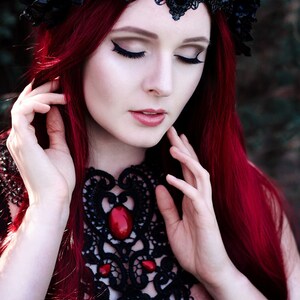 May include: A woman with long, bright red hair wears a black lace choker with red gemstone accents and a black lace crown with a large black gemstone. She has her eyes closed and is touching her hair with both hands.