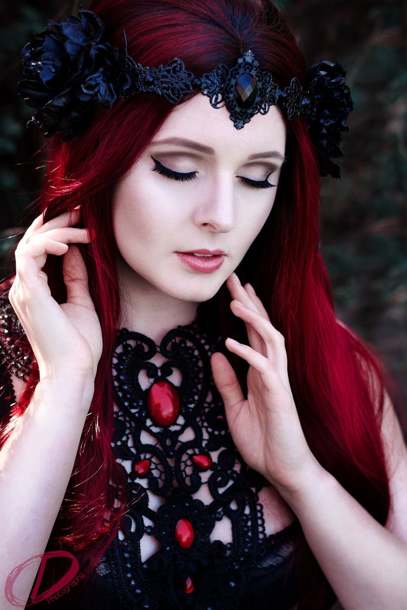 May include: A woman with long, bright red hair wears a black lace choker with red gemstone accents and a black lace crown with a large black gemstone. She has her eyes closed and is touching her hair with both hands.