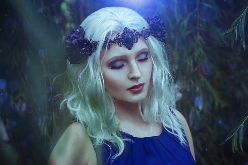 May include: A woman with long, white hair wears a black floral crown with a dark gemstone in the center. She is wearing a dark blue sleeveless dress and has her eyes closed. The background is a blurred image of green foliage.