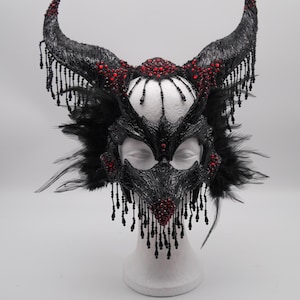 Könnte beinhalten: Eine schwarze Maskenballmaske mit Hörnern, Federn und roten Strasssteinen. Die Maske befindet sich auf einem weißen Mannequin-Kopf.