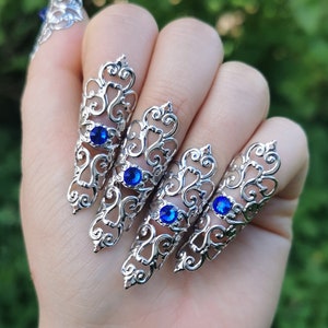Könnte beinhalten: Ein Set aus silberfarbenen Fingerpanzerringen mit einem komplizierten Filigran-Schnörkelmuster. Jedes längliche Teil ist mit einem einzelnen runden, leuchtend blauen Edelstein akzentuiert, der in der Mitte des dekorativen Metalldesigns sitzt.