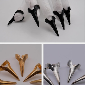 Könnte beinhalten: Eine weiße Hand mit schwarzen, krallenförmigen Fingernägeln. Darunter goldene und silberne Krallenringe. Die Ringe und Fingernägel haben ein ähnliches Design mit einer Spitze und einer gebogenen Basis. Die Ringe sind kreisförmig angeordnet.