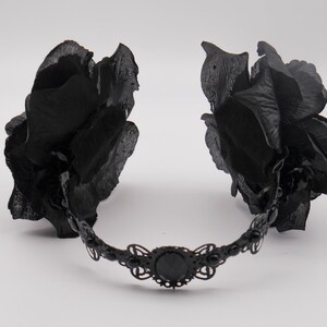 May include: A black headband with two large black fabric roses on either side. The headband is decorated with a black metal design and a black jewel in the center.