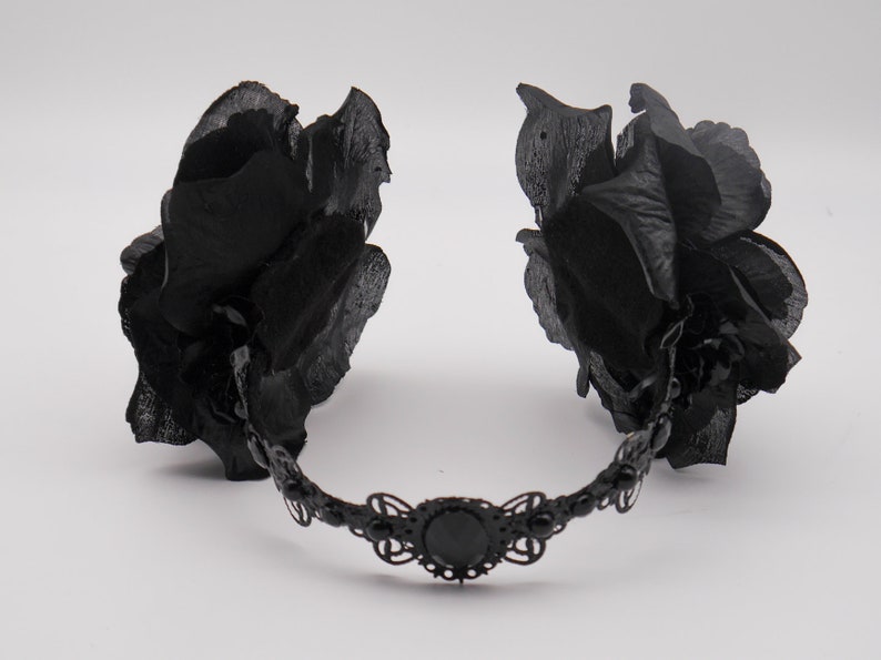 May include: A black headband with two large black fabric roses on either side. The headband is decorated with a black metal design and a black jewel in the center.