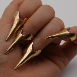 Könnte beinhalten: Goldfarbene Nagelringe im Krallendesign. Die Ringe werden an den Fingern getragen und erzeugen ein scharfes, spitzes Aussehen. Der Schmuck hat ein metallisches Finish und einen einzigartigen, kantigen Stil.