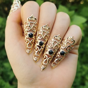 Könnte beinhalten: Vier goldene filigranartige Fingernägelringe mit schwarzen Edelsteinen. Die Ringe sind so konzipiert, dass sie über den Fingernagel passen und eine spitze, klauenartige Form haben.